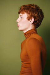 Profile portrait of young man with red hair wearing orange turtleneck sweater against green background in studio. Serious expression and casual style with modern haircut © SHOTPRIME STUDIO