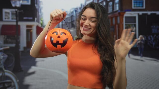 Woman holds jack o lantern bucket in right hand and waves with left hand on a sunny city street; festive happiness.