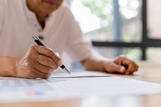 Senior man writing on document at home, close-up of hand holding pen. Retirement planning, financial paperwork, insurance form, legal document, senior lifestyle and careful decision making concept.