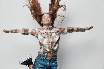 Girl jumping with arms outstretched wearing checkered shirt and denim shorts on white background...