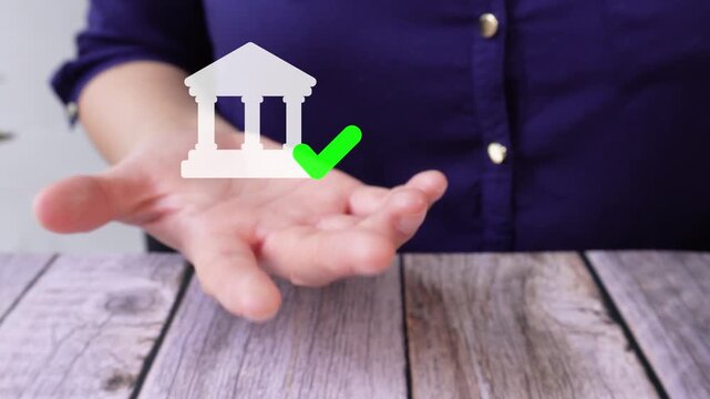 Close-up of a businesswoman's hand on a table holding an animated bank icon with a checkmark