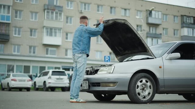 Young white man examines engine compartment in residential parking lot, hood lifted, hands probing components, wearing denim outfit, contemplative expression, silver sedan with slight front damage,