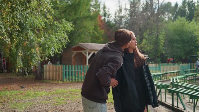 two friends walking along leafy path, discussing layout and pointing at pavilion, green fence and bench rows frame scene, animated gestures and brainstorming energy, soft fall colors and relaxed
