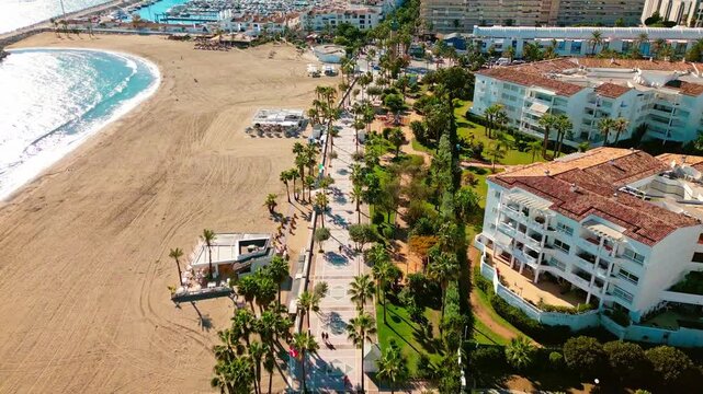 Aerial tilt-up of Puerto Ban&uacute;s beach and harbor, Marbella, Costa del Sol, Spain, featuring terracotta luxury buildings, palm-lined promenade with pedestrians, marina boats, and La Victoria statue