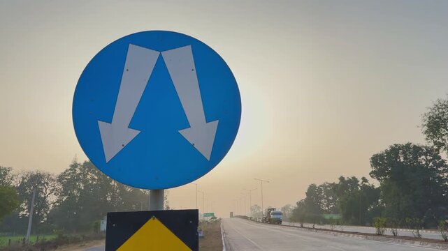 Tracking shot past mandatory pass either side sign on highway, arrows visible as vehicles approach in distance at sunrise with trees lining open roadway ahead calmly now!