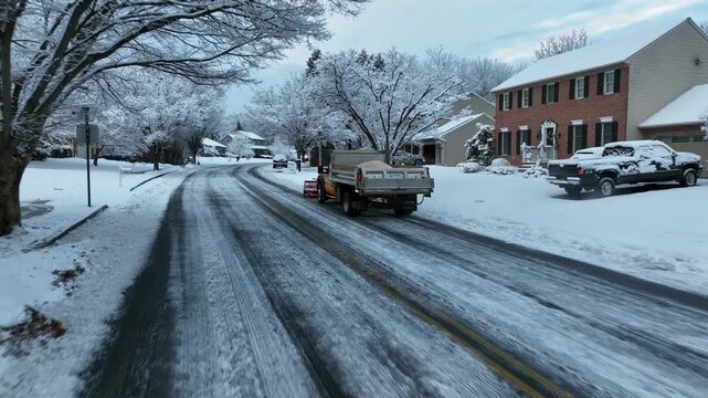 Aerial view of quiet American suburban street after snowfall, with snow-covered houses, icy roads, parked vehicles and frost-covered trees. Snow road salt vehicle cleaning street.