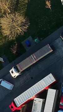 Vertical Screen Aerial Truck Maneuvering Into Parking Slot At Service Bay, Tight Alignment Between Trailers, Close Curb Approach And Careful Navigation On Asphalt Under Late Afternoon Light,