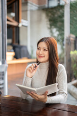 Fototapeta premium Pretty asian woman holding notebook thinking with pen under chin sits at wooden table outside cafe.