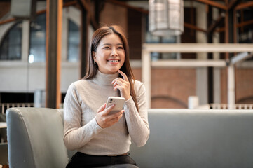 Pretty asian woman holding phone thinking with finger under chin while sitting on sofa couch in cafe
