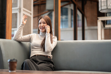 Fototapeta premium Pretty asian woman talking on phone with one hand raised while sitting on sofa in coffeehouse cafe.