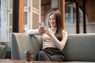 Pretty asian woman holding looking at phone with one elbow on sofa while sitting in coffeehouse cafe