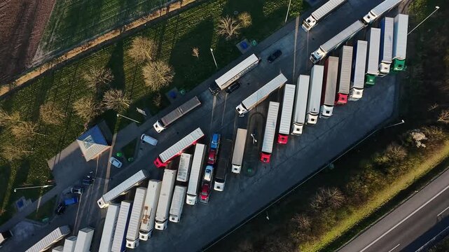 Aerial Truck Parking Lot Filled With Lined Trailers And Idling Engines, Fleet Managers Planning Reroutes, Roadside Pavilion And Highway Visible, Late Afternoon Light And Tense Logistics