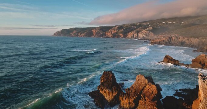 Sintra Cascais Natural Park coastal cliffs stretching along rugged coastline while powerful Atlantic Ocean waves crash against the rocky shore and dramatic sea stacks at sunset. Aerial flight panorama