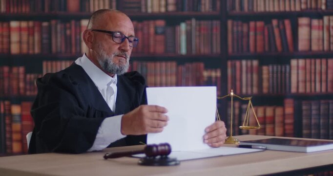 Judge holding legal documents to review in courtroom for justice and judicial decision. Paperwork of busy senior magistrate sitting at desk with law books and balance scales to work on criminal case