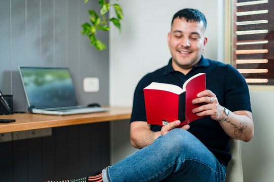 A man in his 30s reads a book in his modern home office with a laptop nearby and plants