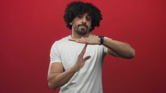 Man with curly hair in white t shirt making timeout gesture with hands and wristwatch in red studio; playful pause timekeeping.