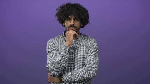 Man with curly hair, hand on chin and crossed arms in studio with purple backdrop, looking at camera; contemplation.