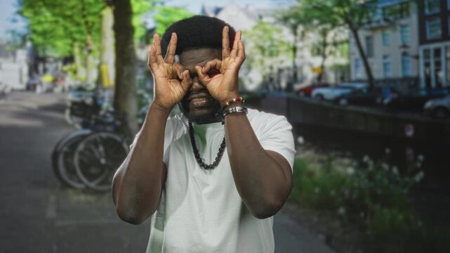 Man making hand binoculars and ok sign on a sunny street by a canal with bicycles and parked cars visible; playful curiosity.