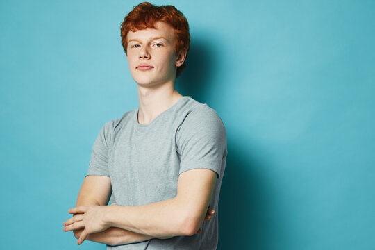 Young redhead man with crossed arms wearing gray t shirt standing confident against blue background looking at camera with relaxed expression portrait
