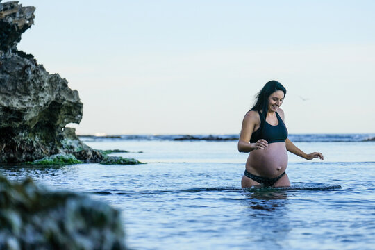 Athletic pregnant woman in black bikini wading in ocean water on calm morning in soft light