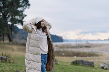 Woman outdoors wearing a long puffer coat in a windy lakeside landscape with fields and trees showing seasonal style and outdoor comfort for travel photos today © SHOTPRIME STUDIO