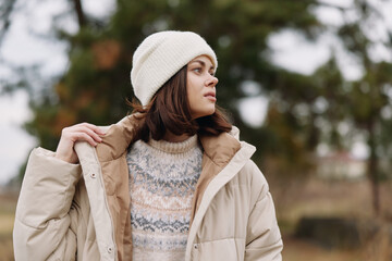 Woman in a beige puffer coat and white beanie stands outdoors, gazing to the side, showcasing winter fashion in a calm, natural portrait amid autumn scenery. © SHOTPRIME STUDIO