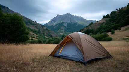 A brown camping tent sits in a dry grassy valley surrounded by rugged mountains under a cloudy sky