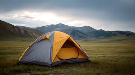 A yellow and grey camping tent is set up in a grassy meadow with rolling hills and snow capped mountains under a dramatic cloudy sky