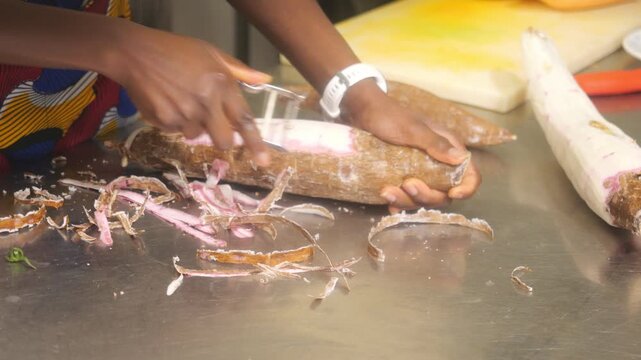 Close up of an African female chef peeling cassava, Manihot esculenta, also known as cassava, yuca, manioc, tapioca, alongside yam, Dioscorea rotundata called igname, &ntilde;ame, inhame, during preparation