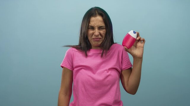 Young hispanic woman grimacing while holding a pink bottle near her face in blue studio wearing a pink t shirt and extended hand; disgust.