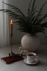 A cup of coffee, a candle in a brass holder, a bouquet in a ceramic vase on a table with a gray...