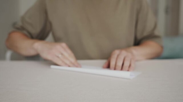 Medium frontal shot of a man folding a sheet of white paper in half on a table. Camera positioned at table height, clear view of the first folding step, calm home atmosphere. High quality 4k footage