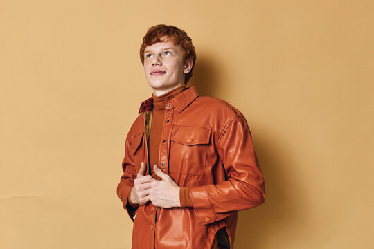 Young man with red hair wearing brown leather jacket standing against beige background. Portrait of confident male model looking up with smile and casual pose in studio light.