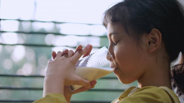 Close up side profile of adorable Asian girl drinking milk from glass inside home near window. Thirsty child holding glass with two hands enjoying healthy dairy nutrition for growth in natural light.