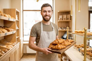 Smiling male baker holding a wooden tray of freshly baked croissants in a bright bakery with shelves of assorted pastries and bread in the background