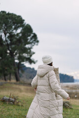 Winter scene of a woman in a long beige puffer coat walking along a lakeside path, knit hat, bare trees and soft light, conveying calm outdoor fashion and serene nature