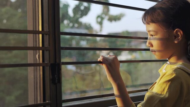Sad Asian girl looking out window through iron bars during sunset. Lonely child waiting with hope, sun shadows on face and reflection on glass. Concept of depression, solitude, or home isolation.
