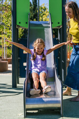 Happy mother watching her young daughter laughing while sliding down a metal slide at a sunny outdoor park © Koldo_Studio