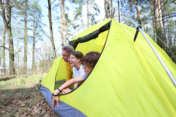 A father and two kids laugh together while peeking from their yellow tent in a sunlit forest. Pure joy, connection, and the magic of family camping adventures.
