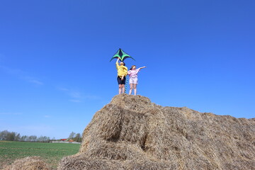 Two kids stand atop a giant haystack under a clear blue sky, launching a vibrant green kite. Their joyful expressions and outstretched arms 