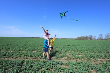 A joyful family moment unfolds in a vast green field under a clear blue sky. A father carries his daughter on his shoulders as she reaches for a soaring green and black kite