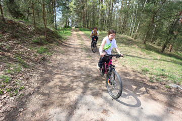 Two kids pedal their bikes along a sunlit forest trail, immersed in natures green embrace. Active, joyful, and carefree childhood adventure at its best.