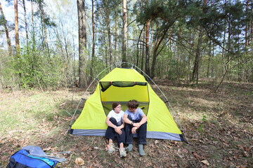Two kids sit side by side at the entrance of their yellow tent in a quiet forest. A peaceful, candid moment of friendship and shared adventure on a sunny camping day.