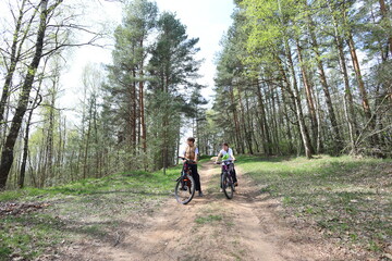 Two kids pause on a forest trail with their bikes, surrounded by tall pines and fresh spring greenery. A moment of rest and connection in natures quiet beauty.