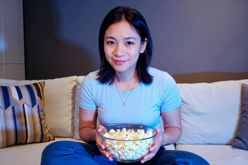 Portrait of young adult Asian woman sitting on sofa holding bowl of popcorn looking into camera smiling, relaxed posture suggesting leisure activity at home