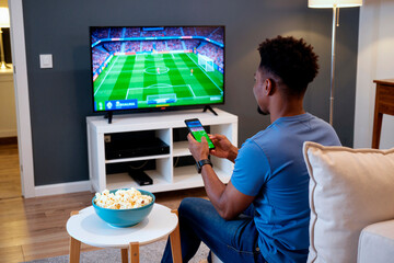 Black young adult man sitting on couch watching soccer match on television while holding smartphone in living room, bowl of popcorn on table in foreground, focused on screen