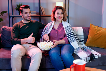 Caucasian young adult man giving thumbs up while sitting next to Caucasian young adult woman eating popcorn on sofa, both smiling and watching television together in living room