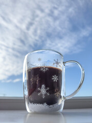 Glass mug with hot black coffee decorated with snowflake pattern against blue sky with copy space