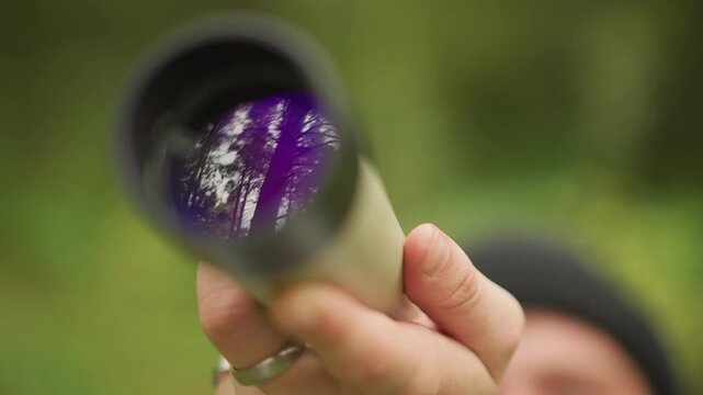 monocular lens showing purple tree reflection held close to camera, hand with rings and beanie hint of face in soft focus, vintage metal tube and green forest backdrop, scout and traveler action