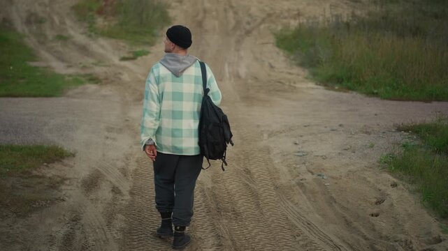 Caucasian hiker dirt crossroads looking back, carrying backpack and plaid jacket, pausing at fork in gravel road, footprints in mud, overgrown grass flanking path, quiet reflective mood during late